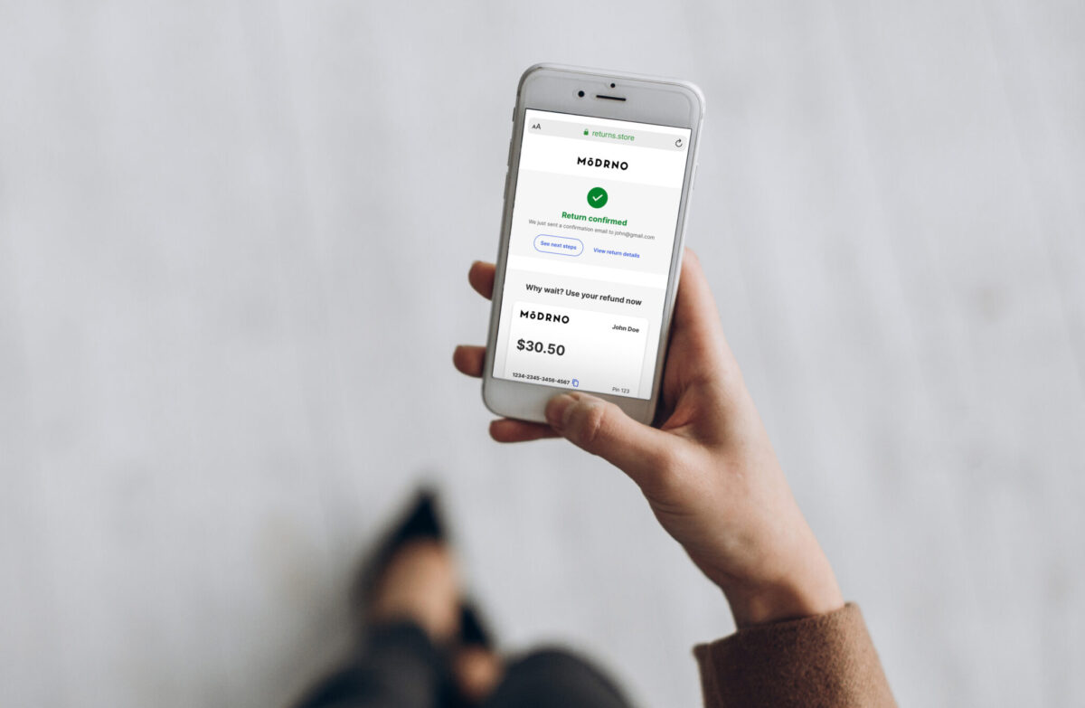 blurred background photo of a professional person wearing black heels holding a mobile phone showing the optoro returns portal with the following imagery and words on the screen: returns.store as the web address, Modrno logo a green checkmark and return confirmed below, we just sent a confirmation email to john@gmail.com, below two buttons that say see next steps and view return details. section below says why wait? use your refund now with a gift card with the modrno logo, John Doe and $30.50 in large letters