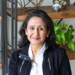 Optoro CEO Amena Ali smiling and sitting in front of a wooden shelving unit with plants and gold accents wearing white button down shirt with black jacket