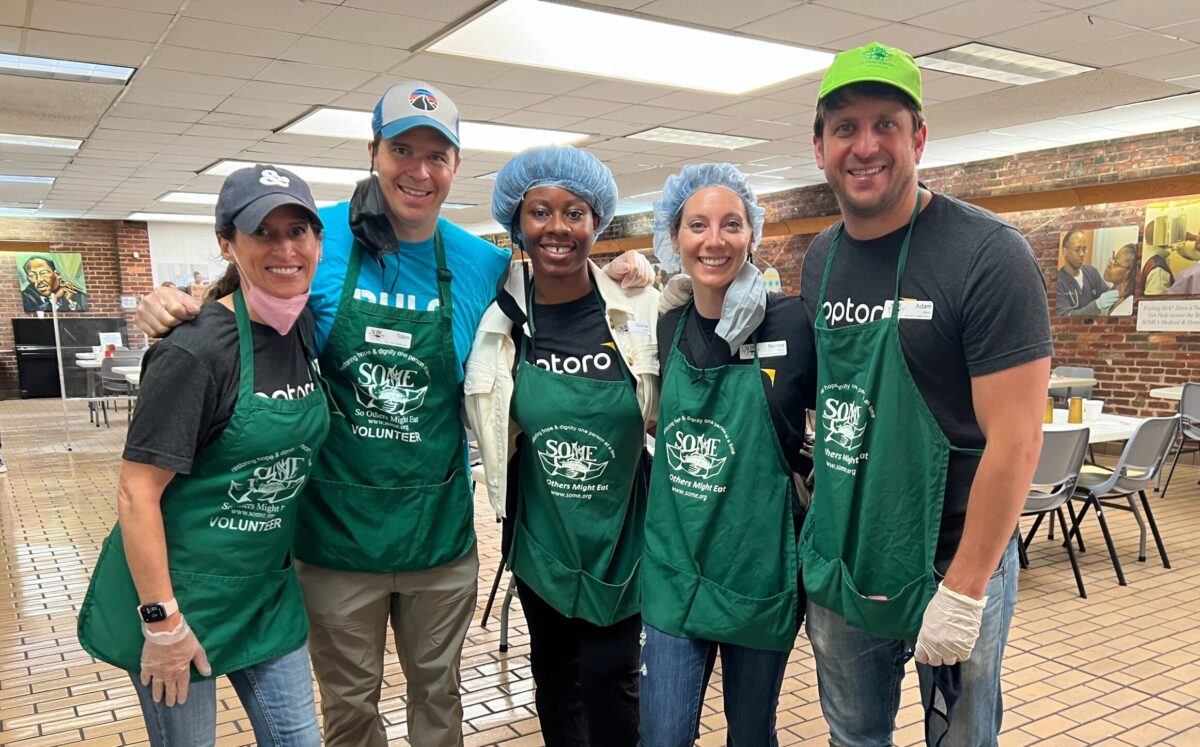 Five Optoro Employees stand smiling with arms around each other in SOME building wearing hair nets or baseball caps and wearing green aprons with SOME.org logo and volunteer printed
