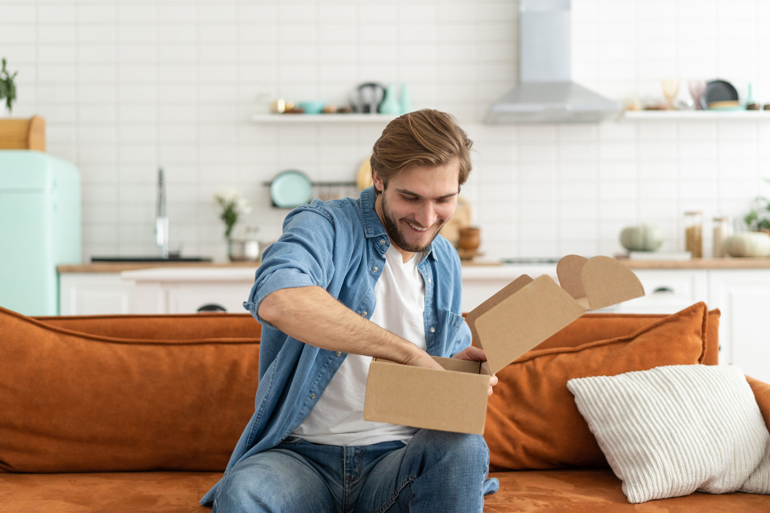 White man with blond hair sits on a burnt orange couch opening a box